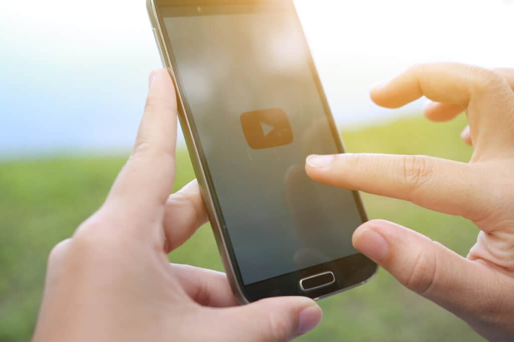 A person is holding a smartphone and about to tap a video play button on the screen. The background is blurred and appears to be outdoors with greenery.